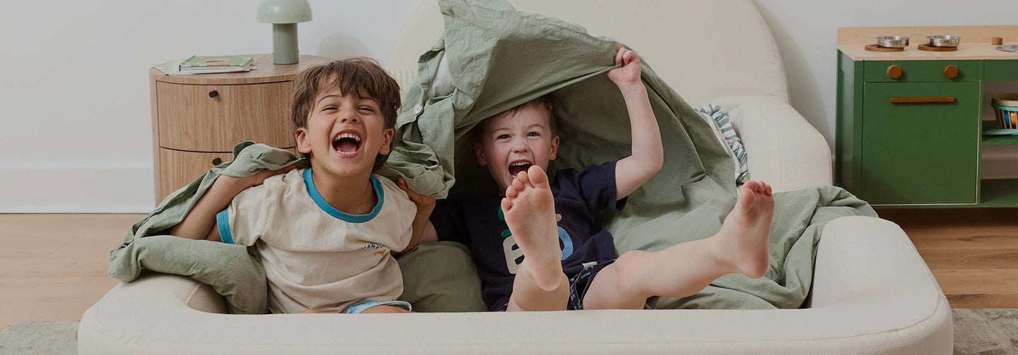 Two children playing under a blanket on a couch in a living room.